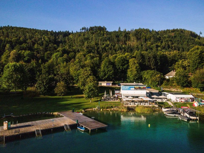 Seeufer mit Steg, Gastgarten und Booten vor bewaldetem Hang in Kärnten